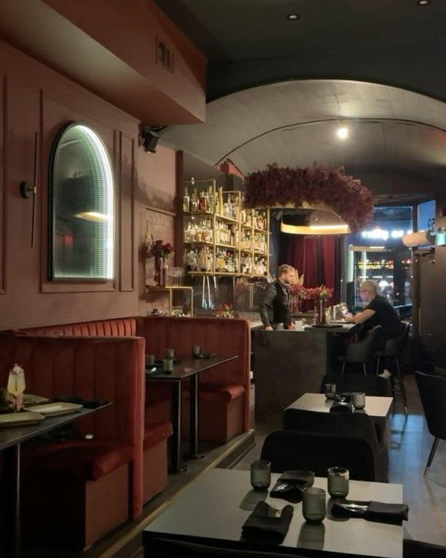 A moody speakeasy dining room with red velvet booths, marble tables and a glowing back bar