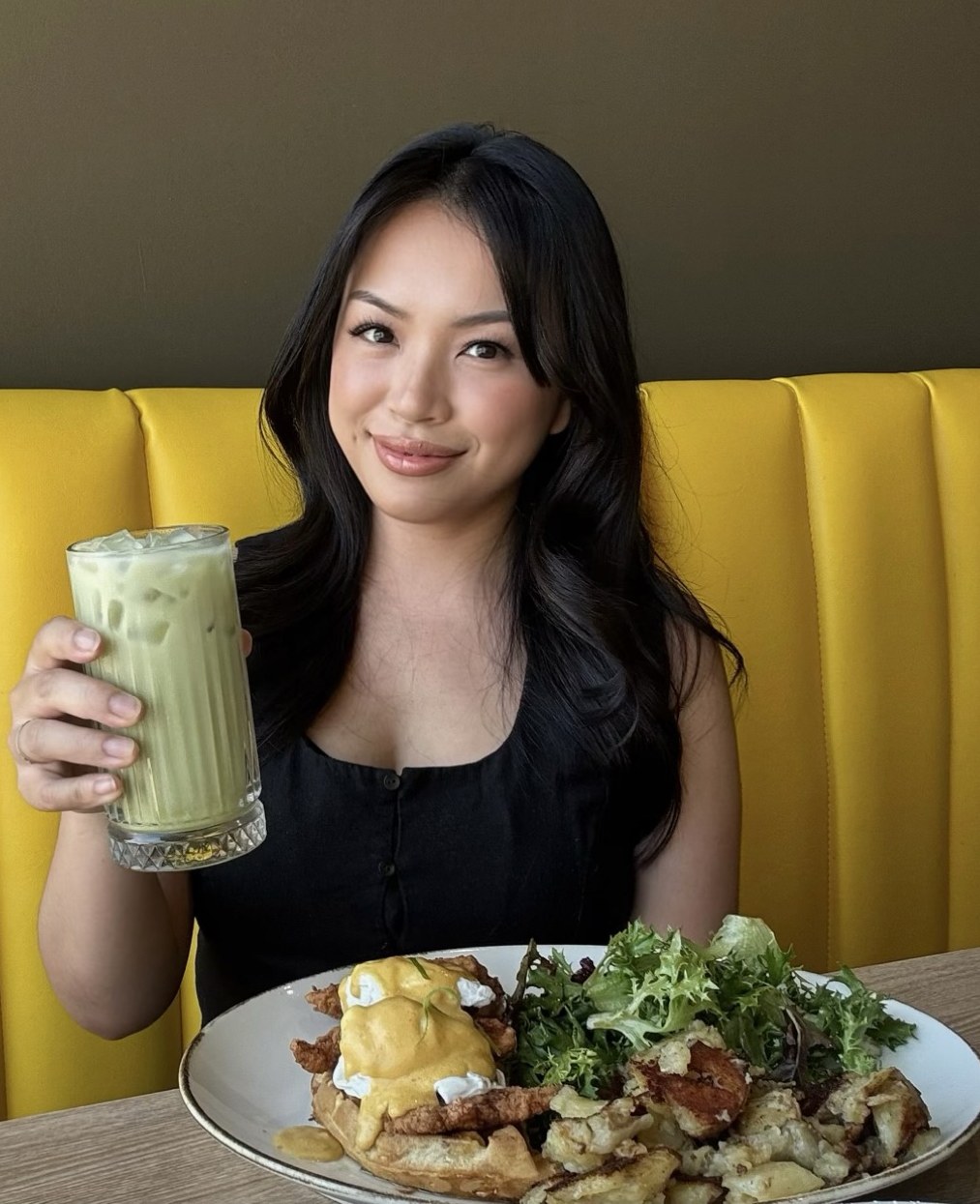Rachel of Brunch Mama holding an iced matcha, seated at a brunch table with eggs benedict